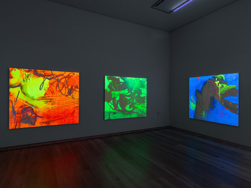 Blick in einen Ausstellungsraum mit Schwarzlicht; an den Wänden hängen neonbunte, abstrakte Gemälde, die im dunklen Raum intensiv leuchten.