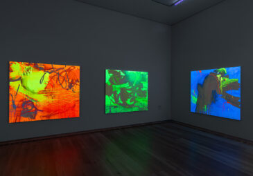 Blick in einen Ausstellungsraum mit Schwarzlicht; an den Wänden hängen neonbunte, abstrakte Gemälde, die im dunklen Raum intensiv leuchten.